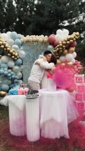 Outdoor gender reveal party with balloons and cake, featuring a couple embracing joyfully.